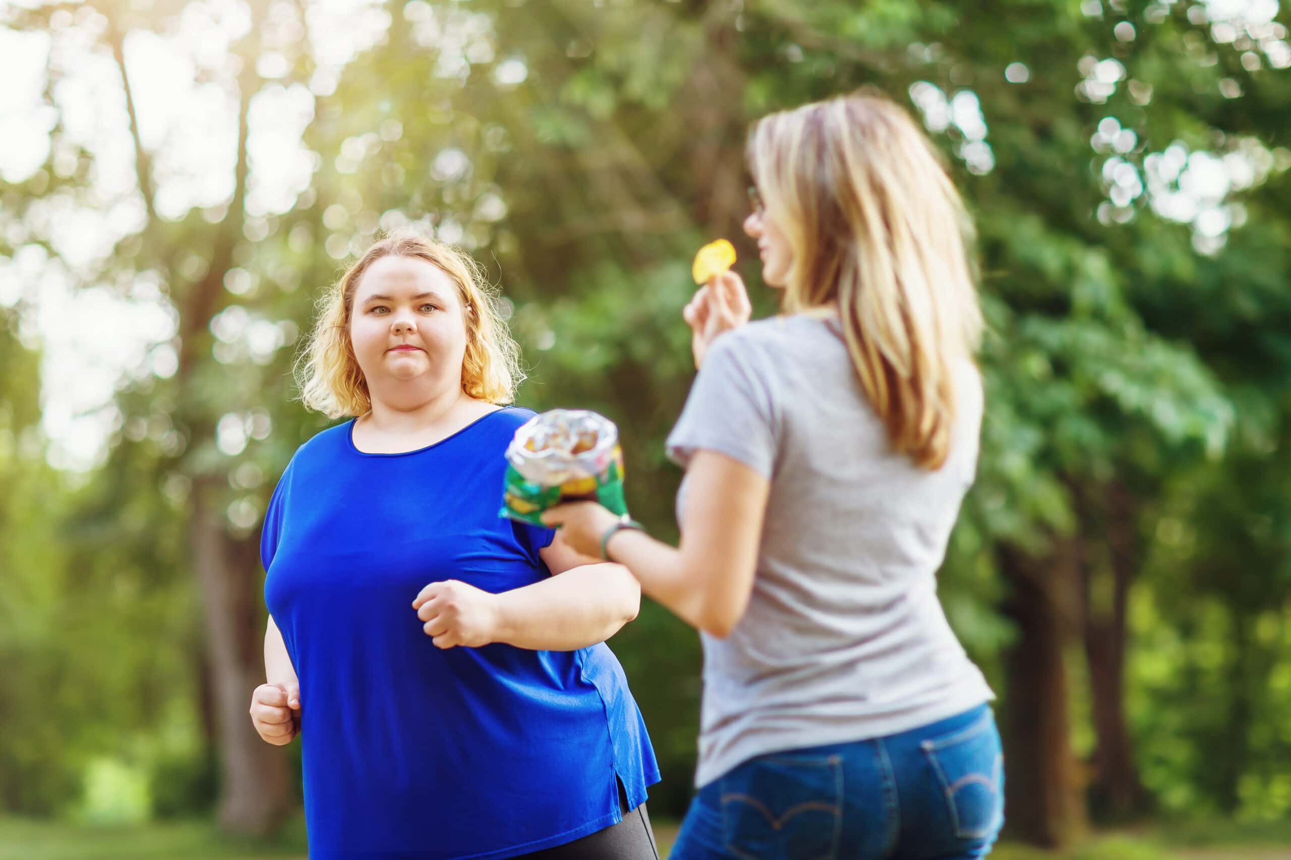 A Young Blonde Of Plus Sizes Runs In The Park Near A Woman Eating Chips. The Concept Of A Healthy Lifestyle And Self Improvement
